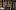 ETC 33 Variations - Ryan Kurtz A woman with short brown hair with an earth-toned shawl stands looking hopeful. A man with white hair in a colonial suit with a blue scarf stands behind her, looking fondly at the woman. Behind them is a hall with walls lined with sheet music and lights, neatly organized into rows.