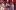Alice in Wonderland-2018-10 A white woman in multi-patterned clothing and a large purple top hat holds a tray of treats. A young white woman in a white turtleneck and blue floral dungarees looks at them suspiciously. Others are gathered watching the exchange.