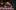 Fortune-2020-12 A man with gray hair stands over a brunette woman with a facial veil as they try to type on a typewriter. They are in a lavishly decorated living room with various knick-knacks on shelves.